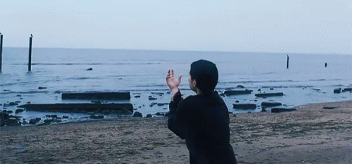 a gif of mitski from the music video for "geyser" mitski is in all black at the beach. she has her right hand in front of her and it is reaching for the ocean but her left hand guides the hand away from the ocean and towards the sand on the left side of the frame. 