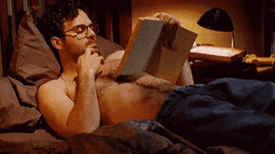 a shirtless man is reading a book while laying in bed .