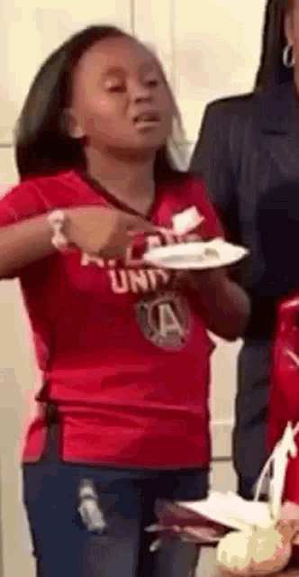 a girl in a red shirt is holding a plate of food and making a funny face .