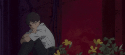 a man in a white shirt sits in front of a red wall with flowers in the foreground