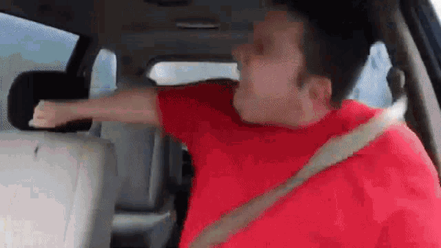 a man in a red shirt is sitting in a car with his arms outstretched .
