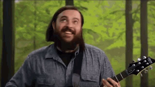 a man with a beard is playing a guitar in front of trees .
