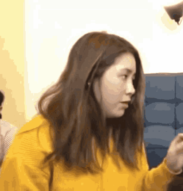 a woman in a yellow sweater is standing in front of a blue couch and looking at something .