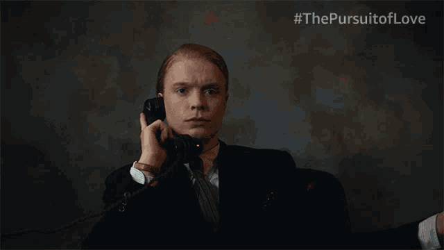 a man in a suit and tie is talking on a phone with #thepursuitoflove written on the bottom right