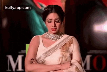 a woman in a white saree and necklace is sitting with her arms crossed .