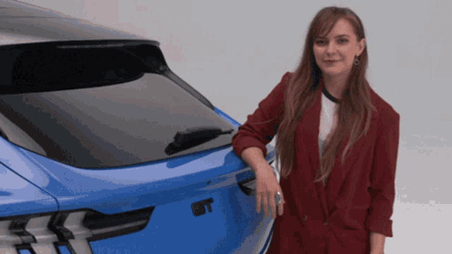 a woman in a red jacket leans against a blue car that says gt