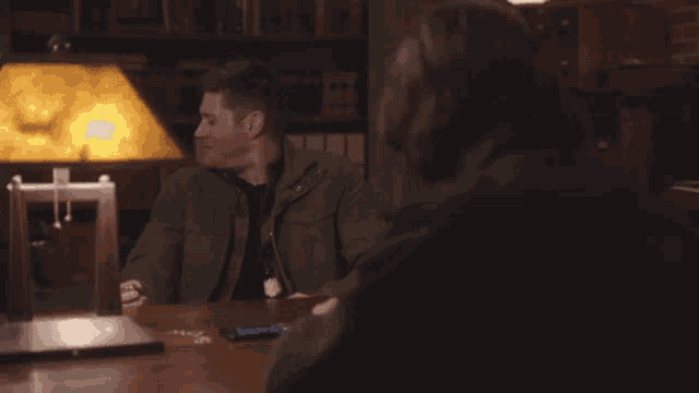 two men are sitting at a table with a lamp and a laptop .