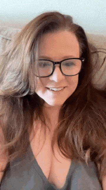 a woman wearing glasses and a gray tank top is smiling while sitting on a couch .