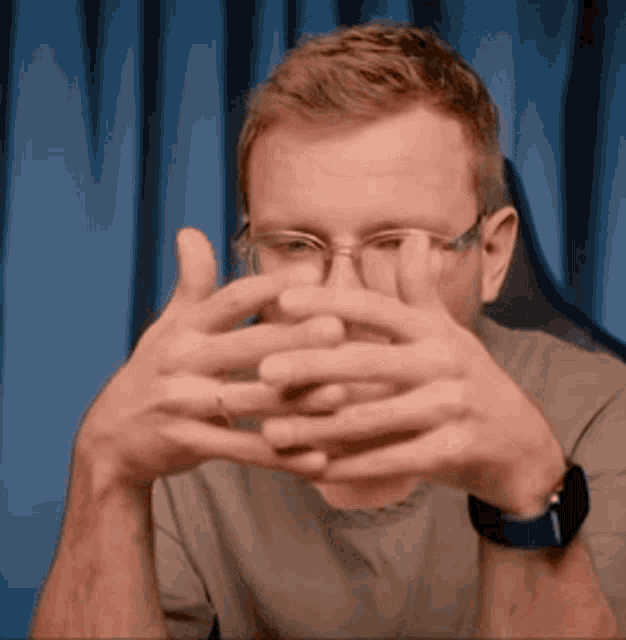 a man with glasses and a watch is covering his face with his hands .