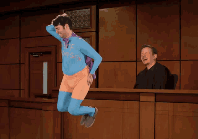 a man in a superhero costume is jumping in the air while another man sits at a counter