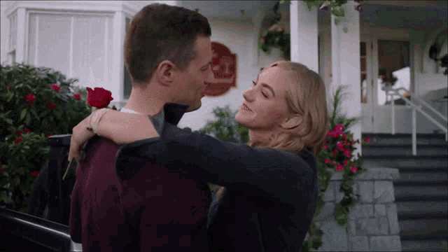 a man is holding a red rose and a woman is hugging him in front of a house