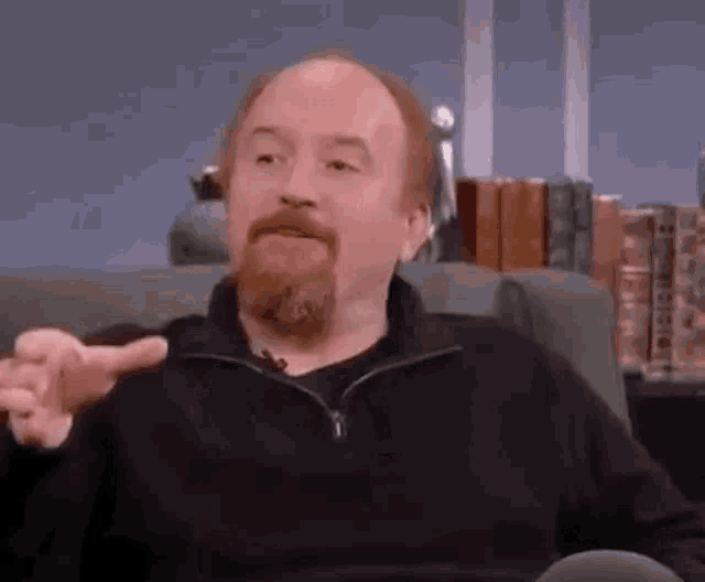 a man with a beard is sitting on a couch and pointing at the camera .