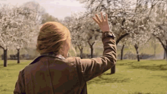 a woman in a brown jacket is standing in a field with her hand up