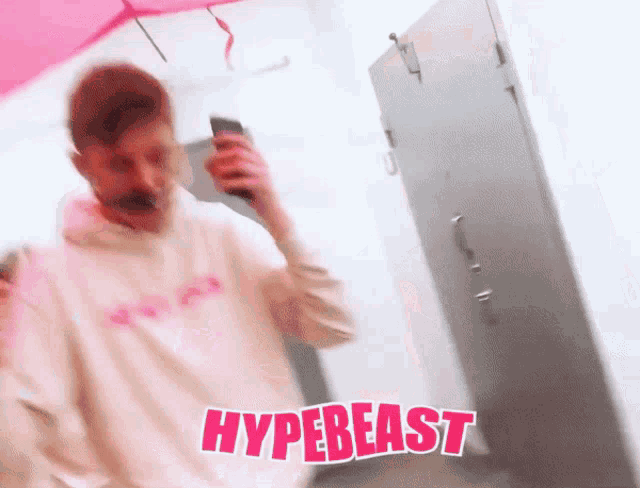 a man in a white hoodie is holding a cell phone and the word hypebeast is on the bottom