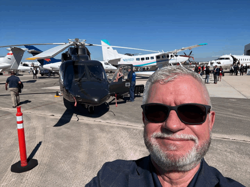 a man wearing sunglasses takes a selfie in front of a helicopter that says navyblue on the side