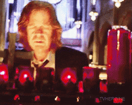 a man in a suit and tie stands in front of red candles with the words tvheroine below him
