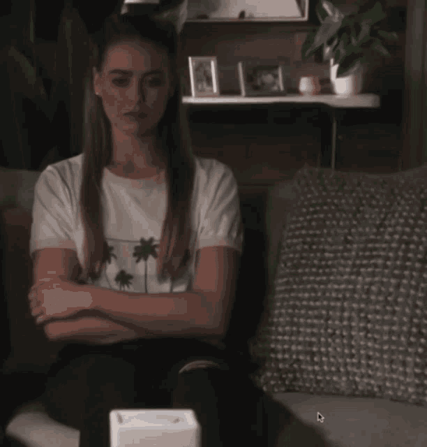 a woman in a white shirt with palm trees on it sits on a couch with her arms crossed