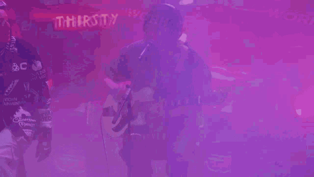 a man is singing into a microphone while another man plays a guitar in front of a purple light .