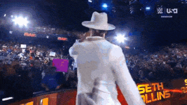 a man in a white suit and hat is standing in front of a crowd at a wrestling event .