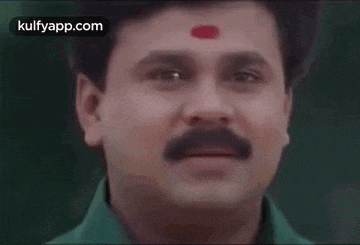 a man with a mustache and a red dot on his forehead is smiling and looking at the camera .