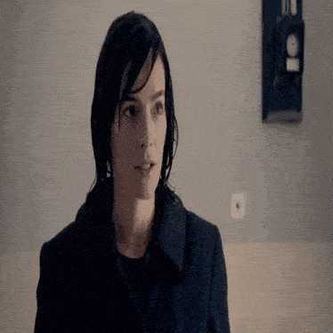 a woman in a black coat stands in front of a wall with a black clock on it