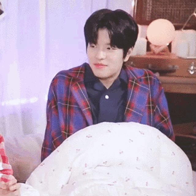 a young man in a plaid jacket is sitting on a bed with a white blanket .