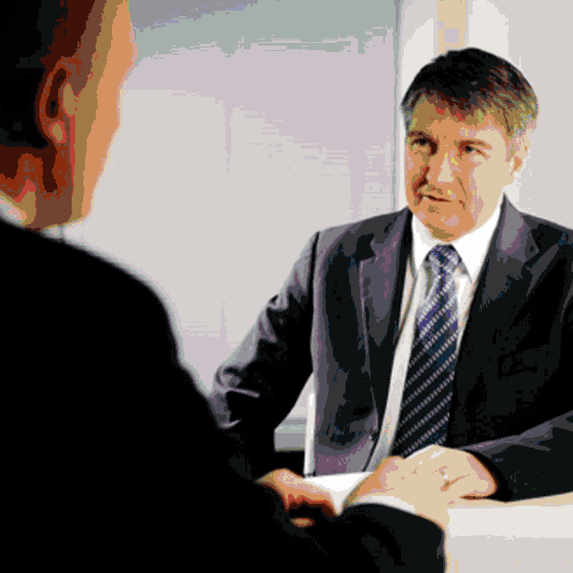 a man in a suit and tie is sitting at a table talking to another man