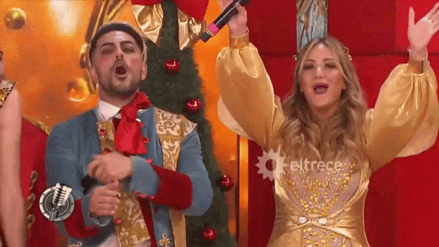a man and woman are singing into a microphone in front of a christmas tree