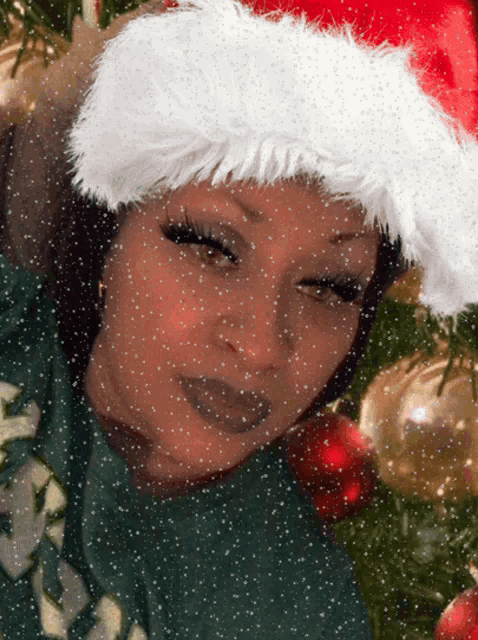 a woman wearing a santa hat stands in front of a snowy christmas tree