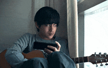 a young man is playing a guitar while looking at a cell phone