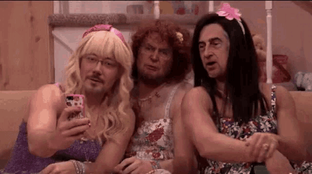 three men dressed as women are sitting on a couch taking a selfie with their cell phones .