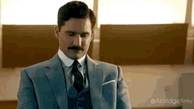 a man in a suit and tie with a mustache is sitting in a room .