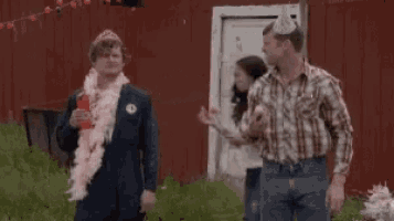a man in a party hat is giving a piece of cake to another man in a pink boa .