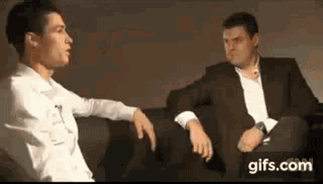 two men are sitting next to each other on a couch talking to each other .
