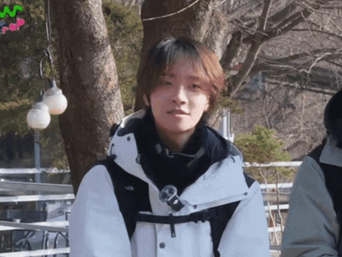 a person wearing a white jacket and a black scarf stands in front of a tree