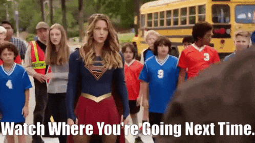 a woman in a superman costume is standing in front of a group of children and a school bus .