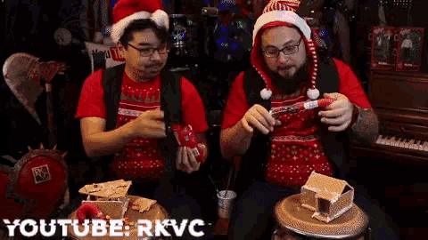 two men wearing santa hats and ugly christmas sweaters are playing with gingerbread houses