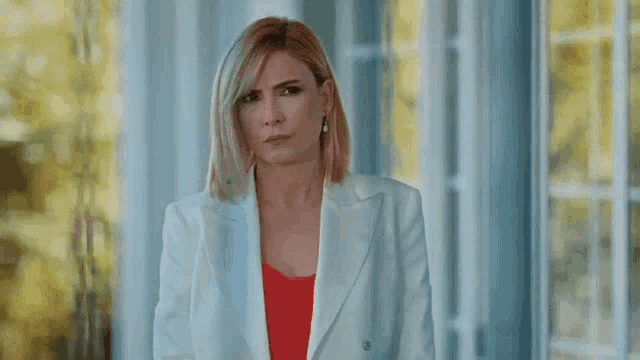 a woman in a white jacket and red top is standing in front of a window looking at the camera .