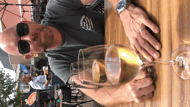 a man wearing sunglasses sits at a table with a glass of wine