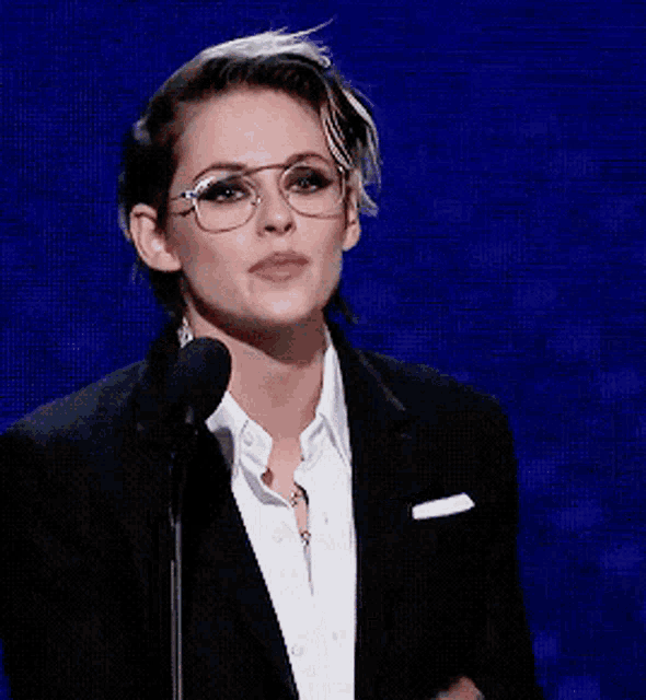 a woman wearing glasses and a tuxedo is speaking into a microphone