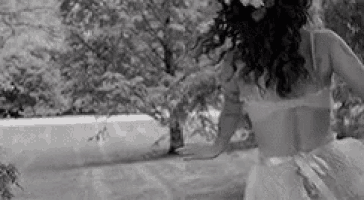 a black and white photo of a woman in a wedding dress standing next to a tree .