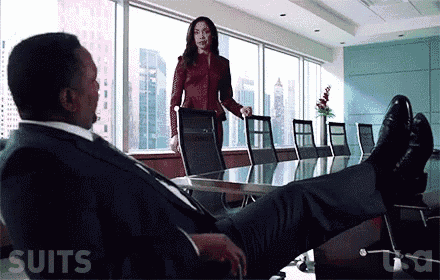 a man in a suit is sitting at a conference table with his feet up and a woman standing behind him .
