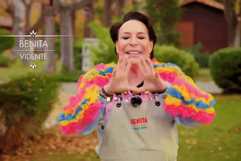 a woman wearing a colorful sweater and an apron that says benita visible makes a heart shape with her hands