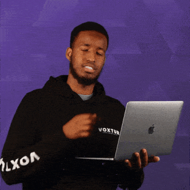 a man wearing a black hoodie that says voxtur on the sleeves is holding an apple laptop