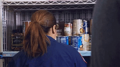 a woman in a blue uniform is standing in front of a shelf full of cans .