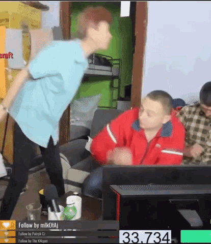 a man in a red jacket is dancing in front of a computer monitor while another man sits on a couch ..