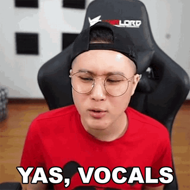 a man wearing glasses and a baseball cap is sitting in a chair and saying yas vocals .
