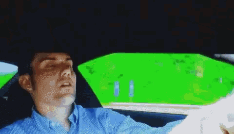 a man in a blue shirt driving a car with a green screen behind him