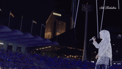a woman in a white dress is singing into a microphone in front of a stadium that says sin herbalife