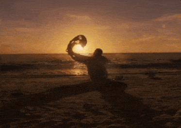 a man is kneeling on the beach at sunset holding a hat in the air .